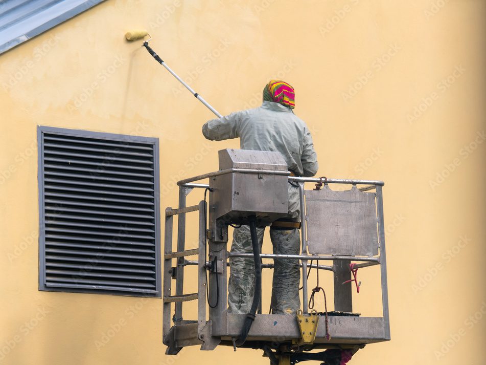 Man on a lifting platform painting the building wall with a roller exterior outdoors. Worker on a ladder manually painting yellow wall on construction site
