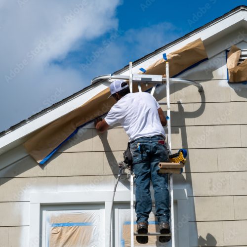 Painter on a ladder masking off trim at the peak of a house before spraying paint